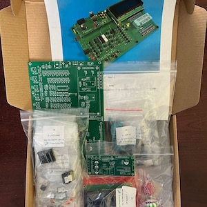 Op de afbeelding: Een open kartonnen doos gevuld met verschillende componenten voor een retro chip-testerkit. De doos bevat verschillende doorzichtige plastic zakken gevuld met kleine elektronische componenten, een groene printplaat en een klein LCD-scherm. De doos bevat ook een gedrukt instructievel met de titel "Retro Chip Tester Professional" en de tekst "Rev. 1.2".