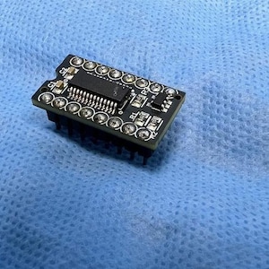 Puede incluir: Una pequeña placa de circuito electrónico negra con múltiples componentes plateados y un chip rectangular central. La placa es rectangular con esquinas redondeadas, sobre un fondo azul.