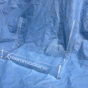 Peut inclure: Un présentoir en acrylique transparent avec le mot "Commodore" gravé sur le devant. Le présentoir est conçu pour contenir un petit objet et présente un design simple et minimaliste. Le présentoir est sur un fond bleu.