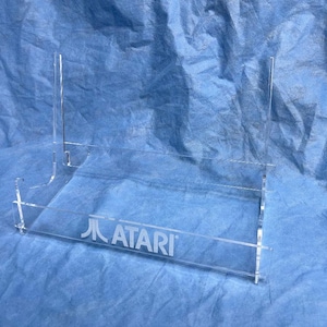 Könnte beinhalten: Transparenter Acryl-Ausstellungsständer mit dem Atari-Logo. Der Ständer hat eine rechteckige Basis und zwei vertikale Stützen. Er ist für die Aufnahme einer Spielkonsole oder anderer Sammlerstücke konzipiert. Der Hintergrund ist ein blauer Stoff.