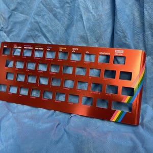 May include: A red-orange keyboard plate with cut-out key spaces and text labels. The plate has a rainbow stripe on the side. Text includes "CAPS LOCK", "TRUE VIDEO", "YELLOW", "GRAPHICS", and "DELETE".