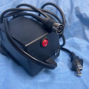 Könnte beinhalten: Schwarzes Netzteil mit rotem Netzschalter und rundem Stecker. Das Netzteil hat ein schwarzes Kabel und einen Standardstecker. Das Gerät befindet sich auf einer blauen Oberfläche.