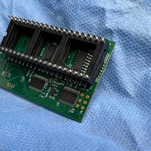 Op de afbeelding: Een groene printplaat met zwarte connectoren en meerdere ge&iuml;ntegreerde circuits. De plaat heeft een rechthoekige vorm met goudkleurige cirkelvormige pads en zilveren pinnen. De achtergrond is een lichtblauwe stof.