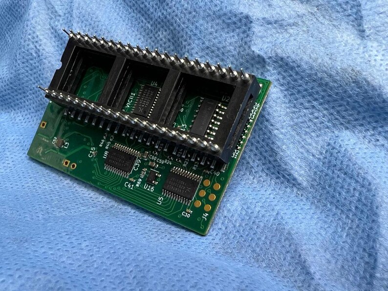 Op de afbeelding: Een groene printplaat met zwarte connectoren en meerdere ge&iuml;ntegreerde circuits. De plaat heeft een rechthoekige vorm met goudkleurige cirkelvormige pads en zilveren pinnen. De achtergrond is een lichtblauwe stof.