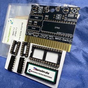 May include: An assortment of electronic components, featuring a black circuit board marked "MAX-Switch" with gold connectors, black sockets, and integrated circuits. The image also includes a label that says "AmericanRetro.shop Commodore, DesTest MAX-Switch".