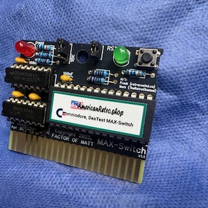 May include: A black circuit board with various electronic components, including resistors and integrated circuits. It features a red and green LED, a button, and the text "AmericanRetro.shop" and "MAX-Switch". The board has gold-coloured pins.