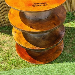 Puede incluir: Una mesa de madera de cuatro niveles hecha con carretes de cable reutilizados. El nivel superior está teñido de naranja y presenta el logotipo de Jack Daniel's y el texto "Tennessee Whiskey".