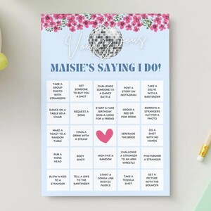 Könnte beinhalten: Eine hellblaue Spielkarte mit dem Text "Maisie's Saying I Do!" und einer Discokugel-Grafik. Die Karte zeigt ein Raster mit lustigen Aktivitäten, mit rosa floralen Akzenten oben. Ein Bleistift, Trauben und ein Notizbuch sind ebenfalls sichtbar.