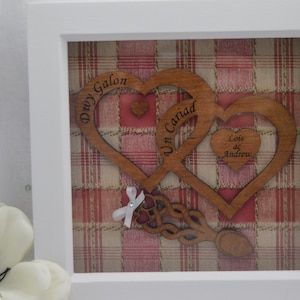 May include: A framed wooden artwork with two intertwined heart shapes, inscribed with the words "Dwy Galon" and "Un Cariad". A smaller heart displays the names "Lois ac Andrew". The backdrop is a red and beige plaid design, all within a white frame.
