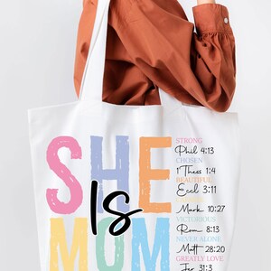 May include: A white tote bag with the words "She is Mom" in colorful letters. The letters are outlined in black and have a rainbow gradient. The bag also has a list of bible verses and words describing a mother, such as "strong", "chosen", "beautiful", "capable", "victorious", "never alone", "greatly love".
