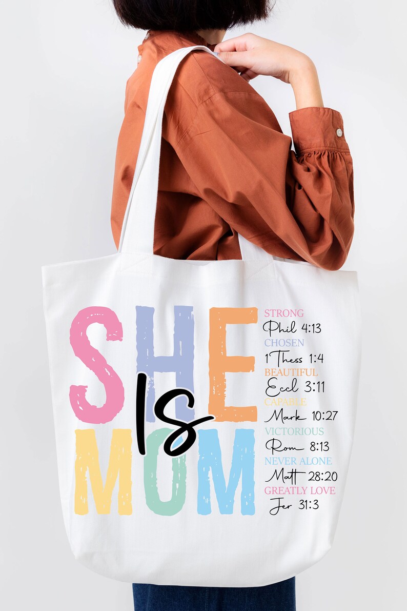 May include: A white tote bag with the words "She is Mom" in colorful letters. The letters are outlined in black and have a rainbow gradient. The bag also has a list of bible verses and words describing a mother, such as "strong", "chosen", "beautiful", "capable", "victorious", "never alone", "greatly love".