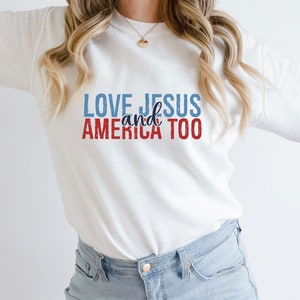 Op de afbeelding: Witte longsleeve sweatshirt met de tekst "LOVE JESUS and AMERICA TOO" in rode, blauwe en witte letters.