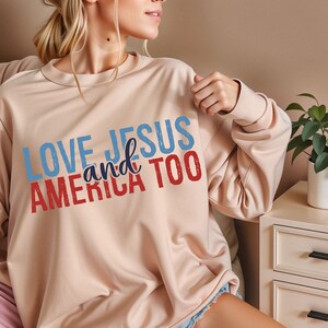 Op de afbeelding: Een lichtbeige sweatshirt met de tekst "LOVE JESUS and AMERICA TOO" gedrukt in rood, wit en blauw.
