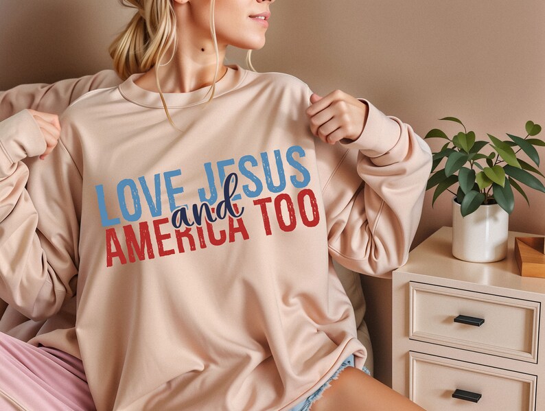 Op de afbeelding: Een lichtbeige sweatshirt met de tekst "LOVE JESUS and AMERICA TOO" gedrukt in rood, wit en blauw.