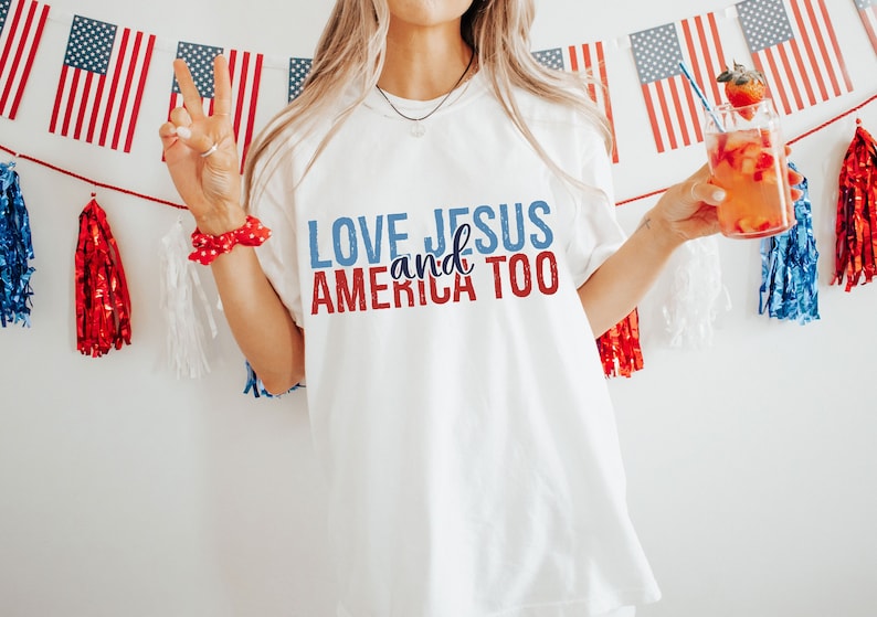 Op de afbeelding: Wit T-shirt met de tekst "LOVE JESUS and AMERICA TOO" in rode, blauwe en witte letters. Het shirt wordt gedragen door een persoon die een glas met rode en oranje vloeistof vasthoudt.
