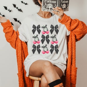 May include: A white t-shirt with a black and pink cherry and bow pattern. The shirt has the text "Spells & Potions" on it.