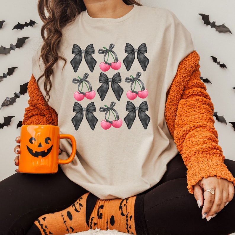 May include: A beige sweatshirt with a black and white graphic print of cherries and bows. The graphic is repeated across the front of the shirt.