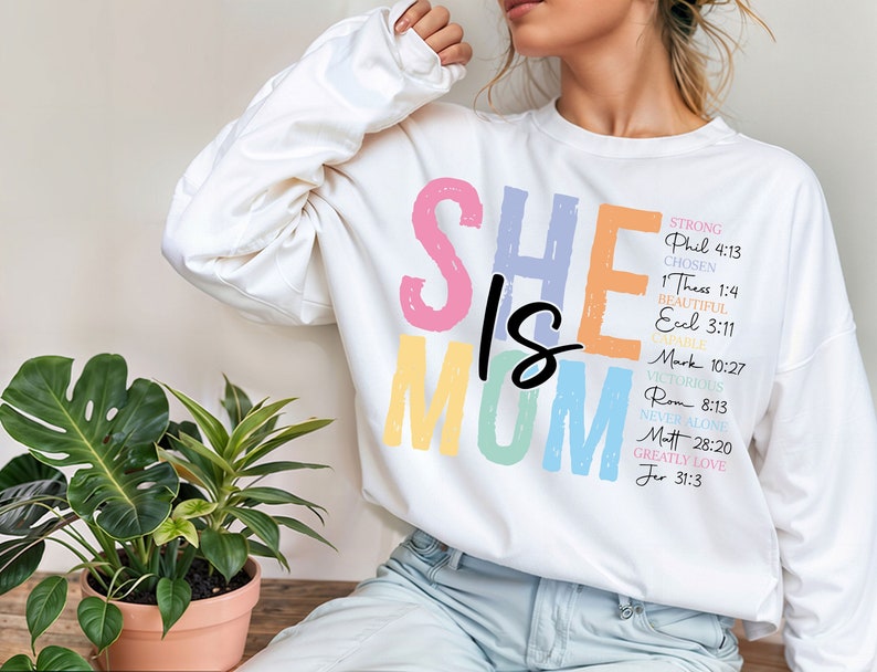 May include: White sweatshirt with the words "She is Mom" in colorful letters. The letters are outlined in black. The text "Strong, Phil 4:13, Chosen, 1 Thess 1:4, Beautiful, Eccl 3:11, Mark 10:27, Victorious, Rom 8:13, Never Alone, Matt 28:20, Greatly Love, Jer 31:3" is printed in black on the right side of the sweatshirt.