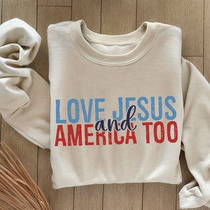 Op de afbeelding: Een beige sweatshirt met de tekst "LOVE JESUS and AMERICA TOO" gedrukt in blauw en rood.
