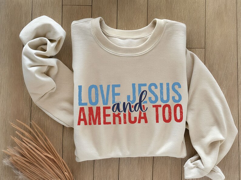 Op de afbeelding: Een beige sweatshirt met de tekst "LOVE JESUS and AMERICA TOO" gedrukt in blauw en rood.
