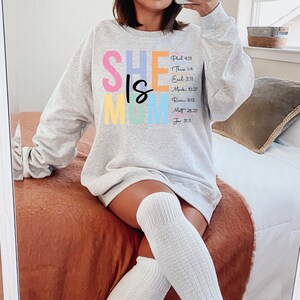 May include: A gray sweatshirt with the text "SHE IS MOM" in colorful letters. The text is accompanied by bible verses.