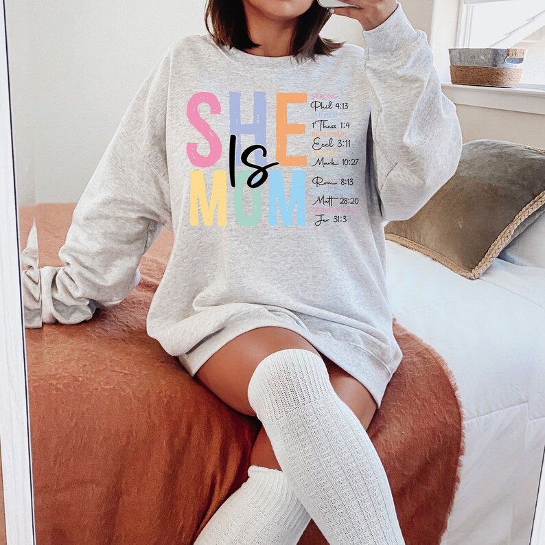 May include: A gray sweatshirt with the text "SHE IS MOM" in colorful letters. The text is accompanied by bible verses.