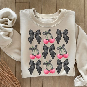 May include: A beige sweatshirt with a black and white cherry and bow pattern. The cherries are pink and the bows are black.