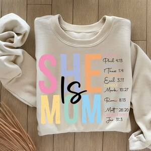 May include: A beige sweatshirt with the words "SHE IS MOM" in colorful letters. The letters are outlined in black and have a slightly distressed look. The text "Phil 4:13, 1 Thess 1:4, Ecel 3:11, CAPAB, Mark 10:27, Rom 8:13, Matt 28:20, GREALLY LOVE, Jer 31:3" is printed in black on the right side of the sweatshirt.