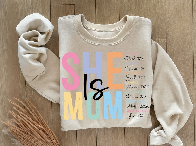 May include: A beige sweatshirt with the words "SHE IS MOM" in colorful letters. The letters are outlined in black and have a slightly distressed look. The text "Phil 4:13, 1 Thess 1:4, Ecel 3:11, CAPAB, Mark 10:27, Rom 8:13, Matt 28:20, GREALLY LOVE, Jer 31:3" is printed in black on the right side of the sweatshirt.