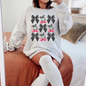 May include: A gray sweatshirt with a black and white print of cherries and bows. The cherries are pink and the bows are black.