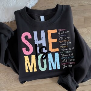 May include: A black sweatshirt with the text "SHE IS MOM" in colorful letters. The text is surrounded by a list of bible verses.