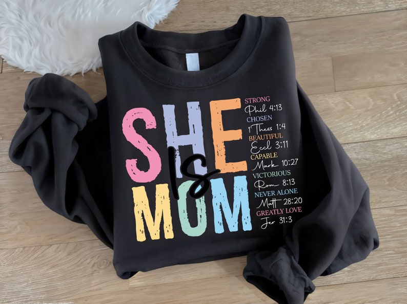 May include: A black sweatshirt with the text "SHE IS MOM" in colorful letters. The text is surrounded by a list of bible verses.