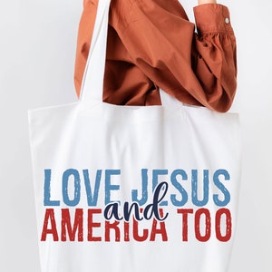 Op de afbeelding: Witte canvas tote bag met de tekst "LOVE JESUS and AMERICA TOO" in rode, blauwe en donkerblauwe letters.