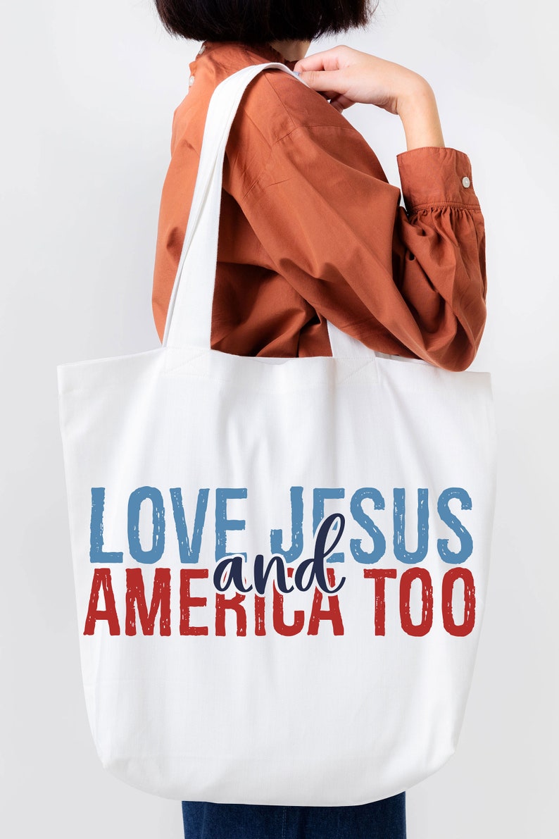 Op de afbeelding: Witte canvas tote bag met de tekst "LOVE JESUS and AMERICA TOO" in rode, blauwe en donkerblauwe letters.