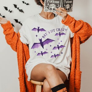 May include: A white t-shirt with the text "Bat Shit Crazy" and purple bats printed on it. The person is wearing an orange cardigan and orange socks with a jack-o-lantern pattern.
