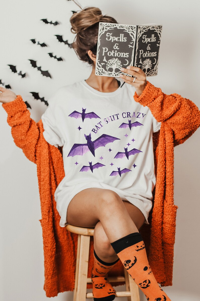 May include: A white t-shirt with the text "Bat Shit Crazy" and purple bats printed on it. The person is wearing an orange cardigan and orange socks with a jack-o-lantern pattern.