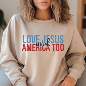 Op de afbeelding: Een beige sweatshirt met de tekst "LOVE JESUS and AMERICA TOO" in rood, blauw en wit.