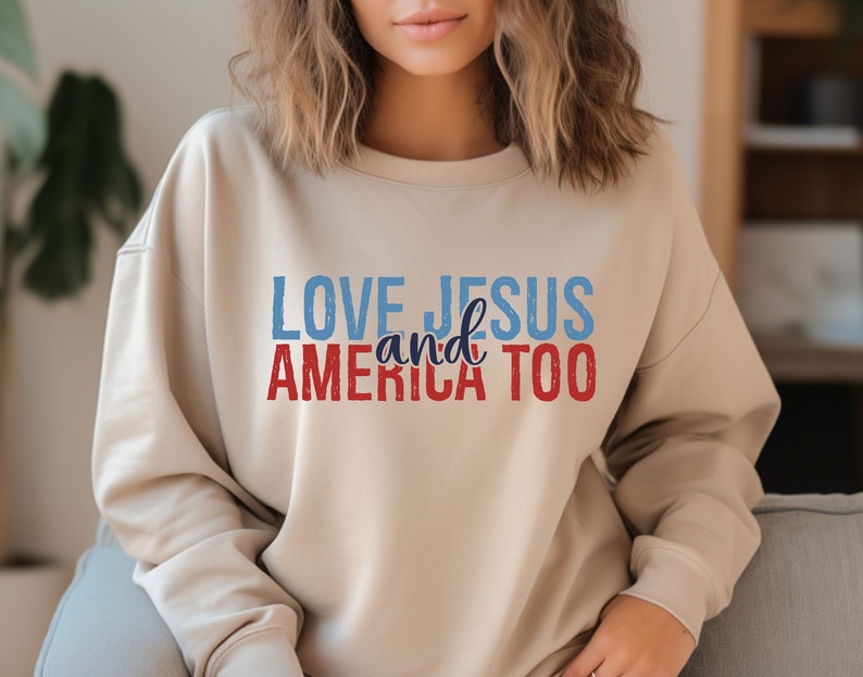 Op de afbeelding: Een beige sweatshirt met de tekst "LOVE JESUS and AMERICA TOO" in rood, blauw en wit.