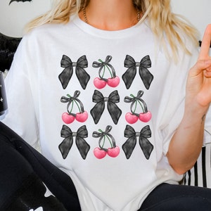 May include: White t-shirt with a black and white graphic print of cherries and bows. The cherries are pink and the bows are black.