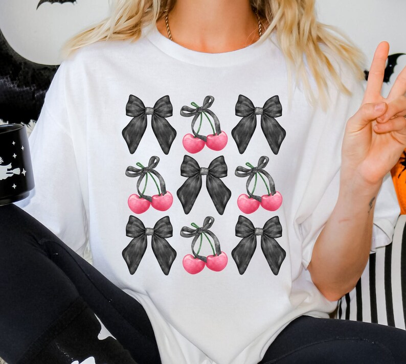 May include: White t-shirt with a black and white graphic print of cherries and bows. The cherries are pink and the bows are black.