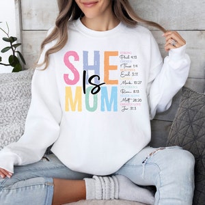 May include: White sweatshirt with the text "SHE IS MOM" in rainbow colors. The text is surrounded by a list of bible verses.