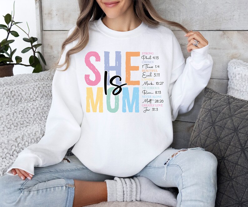 May include: White sweatshirt with the text "SHE IS MOM" in rainbow colors. The text is surrounded by a list of bible verses.