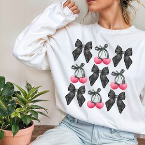May include: White sweatshirt with a black and white print of cherries and bows. The cherries are pink and the bows are black.