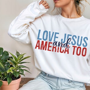Op de afbeelding: Witte longsleeve trui met de tekst "LOVE JESUS and AMERICA TOO" bedrukt in rood, blauw en wit.