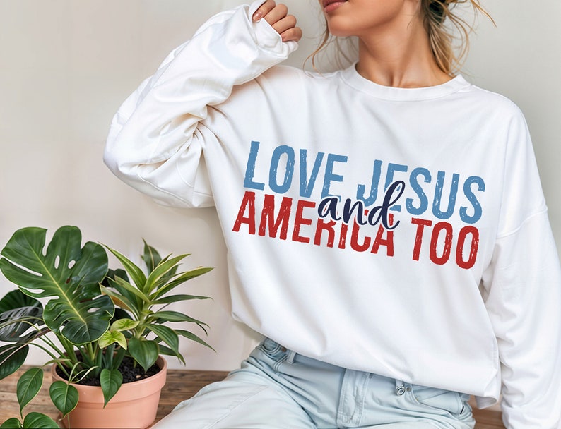 Op de afbeelding: Witte longsleeve trui met de tekst "LOVE JESUS and AMERICA TOO" bedrukt in rood, blauw en wit.