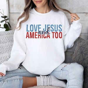 Op de afbeelding: Wit sweatshirt met de tekst "LOVE JESUS and AMERICA TOO" in rode, blauwe en witte letters.