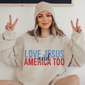 Op de afbeelding: Een beige sweatshirt met de tekst "LOVE JESUS and AMERICA TOO" in blauwe en rode letters.