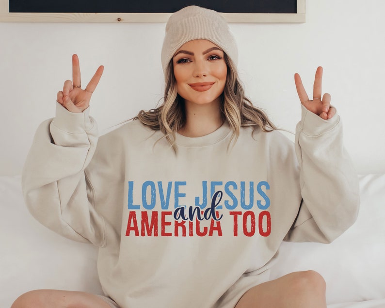 Op de afbeelding: Een beige sweatshirt met de tekst "LOVE JESUS and AMERICA TOO" in blauwe en rode letters.