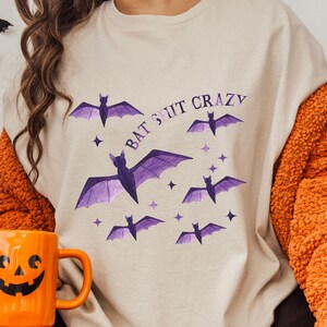 May include: A beige t-shirt with the text "Bat Shit Crazy" printed in purple. The shirt features a watercolor design of purple bats with a white background.