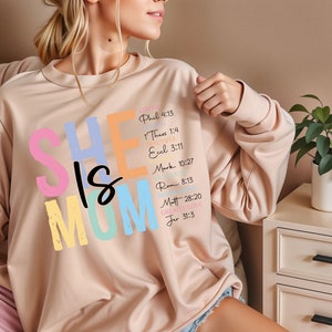May include: A light pink sweatshirt with the text "SHE IS MOM" in colorful letters. The text is surrounded by bible verses in a smaller font.
